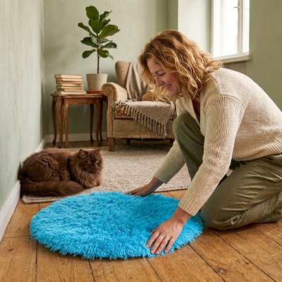 Une femme installe un tapis pour chat rond bleu en fausse fourrure cÎté d'un chat marron