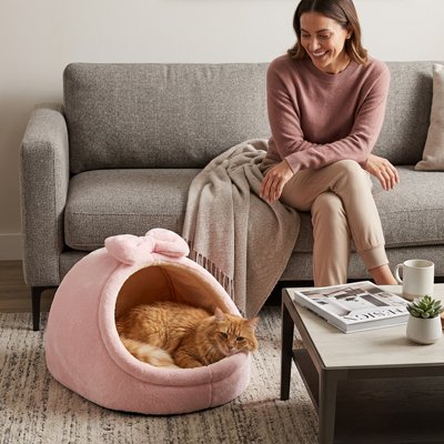 Femme souriante assise sur un canapé à côté de son chat roux installé dans une niche pour chat rose en peluche avec un nœud.