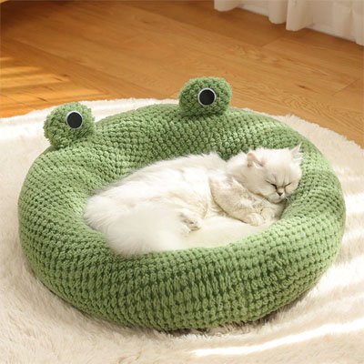 Un chat blanc à poils longs dort paisiblement dans une niche pour chat en peluche verte en forme de grenouille, posé sur un tapis moelleux