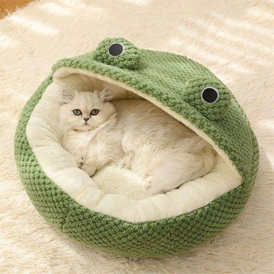Un chat blanc est confortablement allongé dans une niche pour chat en peluche verte en forme de grenouille, posé sur un tapis