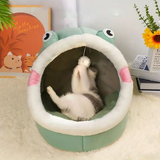 Un chat blanc et gris allongé sur le dos à l'intérieur d'une niche pour chat verte en forme de grenouille, s'amusant avec un pompon suspendu.