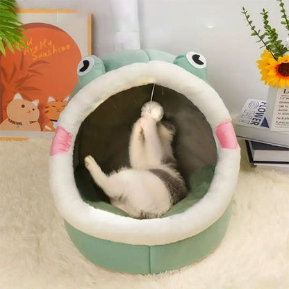 Un chat blanc et gris allongé sur le dos à l'intérieur d'une niche pour chat verte en forme de grenouille, s'amusant avec un pompon suspendu.