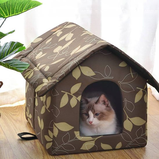 Un chat dans une niche exterieur pour chat en tissu marron à motifs de feuilles jaunâtres, posée sur un parquet en bois