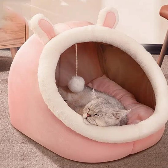Un chaton gris tigré dort paisiblement à l'intérieur d'une niche pour chat en peluche rose avec des petites oreilles de lapin et un pompon suspendu.