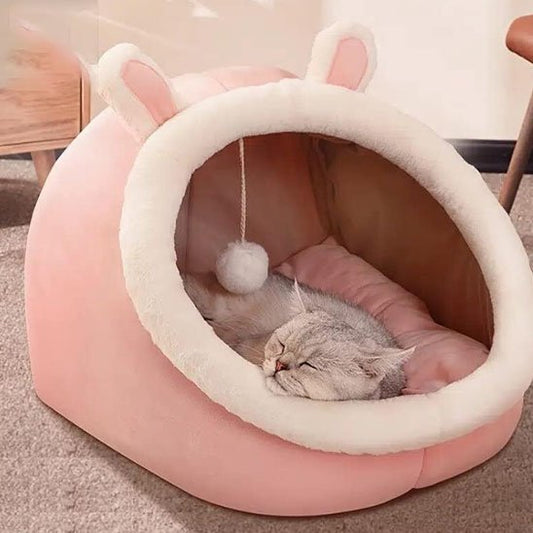 Un chaton gris tigré dort paisiblement à l'intérieur d'une niche pour chat en peluche rose avec des petites oreilles de lapin et un pompon suspendu.