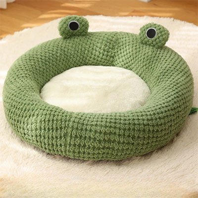 Une niche pour chat en peluche verte en forme de tête de grenouille avec un coussin central crème, posé sur un tapis blanc moelleux